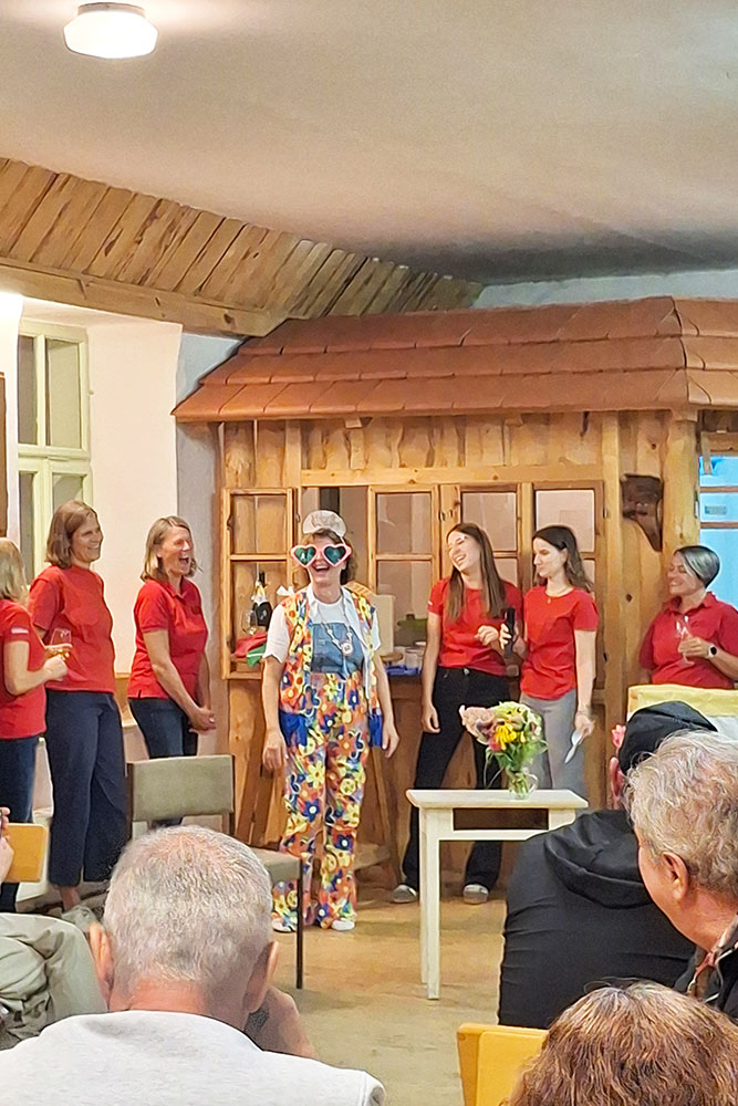 mehrer Personen stehen hinter einer Frau mit Hippioutfit, rosaroter Herzbrille und einer Krone